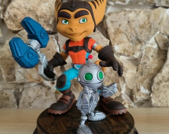 May include: A detailed figurine of Ratchet and Clank, characters from the video game series. Ratchet, with orange fur and blue pants, holds a blue wrench. Clank, a silver robot, stands beside him. The figures are on a wooden base.
