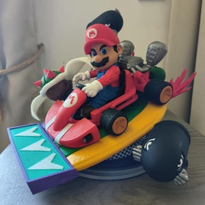 May include: A detailed figurine of Mario driving a red kart with a yellow and blue base. The kart features black tyres, a green and purple front, and a black cannon. Bowser and a Bullet Bill are also included in the design.