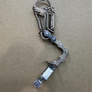 Könnte beinhalten: Ein handgefertigter Metall-Schlüsselanhänger mit einem einzigartigen Design. Der Schlüsselanhänger enthält eine Fahrradkette, eine Feder und einen Verschluss. Das Metall ist silberfarben, und das Gesamtdesign ist industriell und künstlerisch.