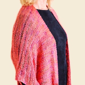 May include: A pink and red knitted shawl with a lacy pattern. The shawl is draped over a person's shoulders and is worn with a black top.