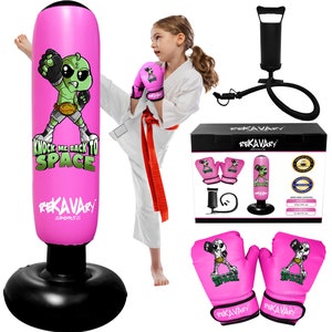 May include: Pink inflatable punching bag with a cartoon alien graphic and the text "Knock Me Back To Space". The bag is on a black base. A girl in a white karate uniform is kicking the bag. Two pink boxing gloves with the same alien graphic are included. The set also includes a black hand pump and a hose.