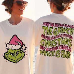 May include: Cream-coloured t-shirt with a Grinch graphic on the front and text on the back. The Grinch has a green face, a pink Santa hat, and stars. The back of the shirt has the text: "JUST SO WE'RE CLEAR THE GRINCH NEVER REALLY HATED CHRISTMAS HE HATED PEOPLE WHICH IS FAIR."