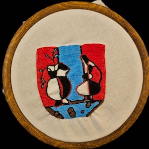 May include: Embroidered artwork in a wooden hoop. The design features two stylized figures in black, white, and red against a blue and red background. The artwork is on a cream-colored fabric.