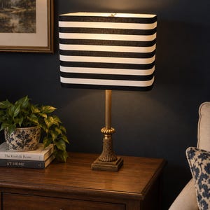 May include: A table lamp featuring a rectangular lampshade with horizontal black and white stripes. The lamp has a slender, metallic gold-toned base with a textured, flared pedestal and a square foot, providing a stable foundation for the light.