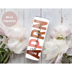 May include: A white 20 oz. tumbler with the name "Jennifer" written in black script and the word "APRN" in large, colorful letters. The letters are in shades of brown, orange, and pink. The tumbler is sitting on a white wooden surface with white flowers.