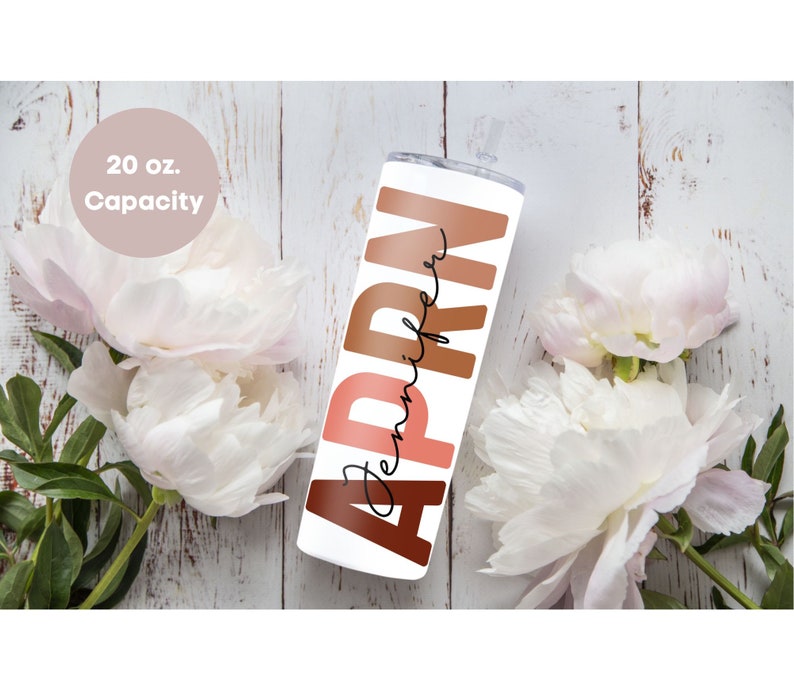 May include: A white 20 oz. tumbler with the name "Jennifer" written in black script and the word "APRN" in large, colorful letters. The letters are in shades of brown, orange, and pink. The tumbler is sitting on a white wooden surface with white flowers.