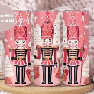 May include: Three pink stainless steel tumblers with a Christmas nutcracker design. Each tumbler features a nutcracker in a red and black uniform, standing in front of a white Christmas tree. The background is a pink and white pattern with snowflakes.