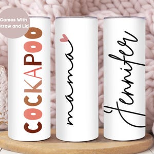 May include: Three white insulated tumblers. One tumbler has the word "COCKAPOO" in shades of brown and pink. Another has the word "mama" with a pink heart. The third tumbler has the name "Jennifer" in black script. All tumblers have lids and straws.