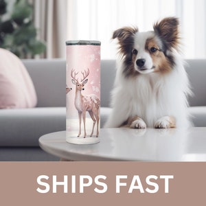 May include: A pink and white stainless steel tumbler with a design of two deer in a snowy forest. The tumbler is on a white table with a dog sitting on the table.