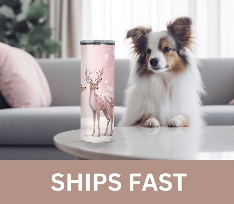 May include: A pink and white stainless steel tumbler with a design of two deer in a snowy forest. The tumbler is on a white table with a dog sitting on the table.