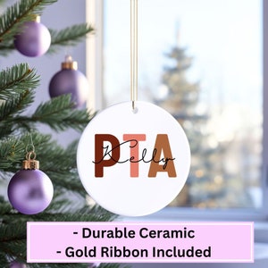 May include: A white ceramic Christmas ornament with the letters PTA in brown, pink, and orange. The name "Kelly" is written in cursive below the letters. A gold ribbon is attached to the ornament.