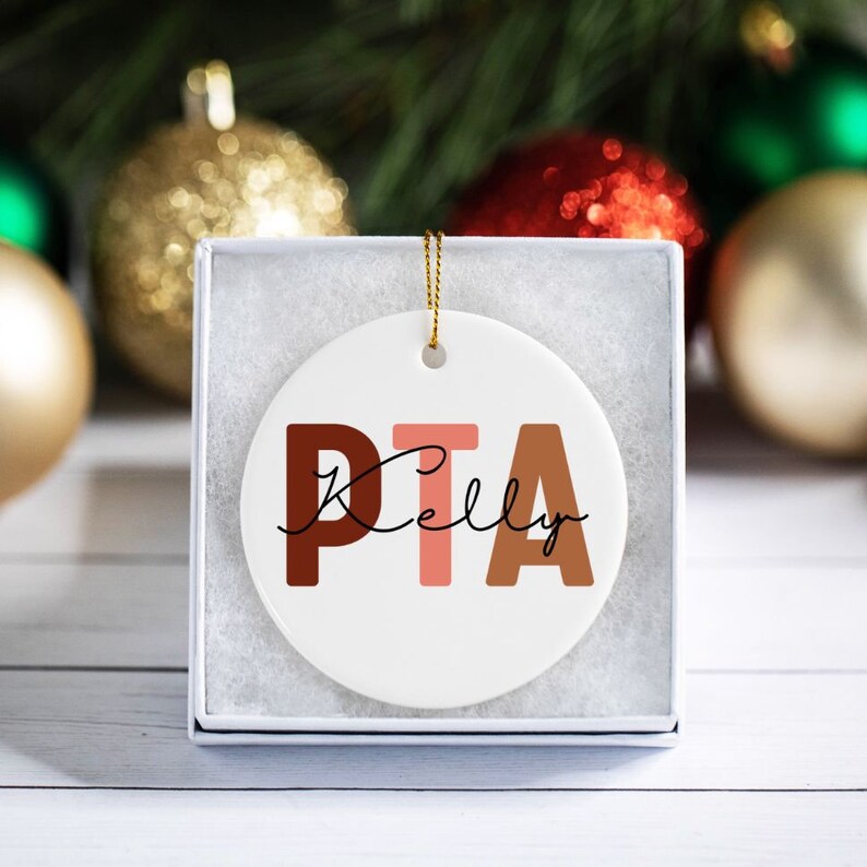 May include: A white ceramic ornament with the letters "PTA" in brown, pink, and orange. The name "Kelly" is written in cursive below the letters.