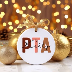 May include: White ceramic ornament with the text "PTA Kelly" in brown, pink, and beige letters. The ornament is surrounded by gold Christmas ornaments.