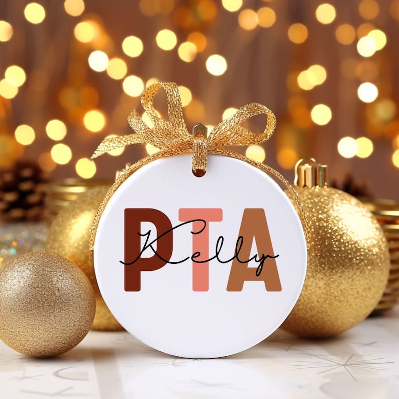 May include: White ceramic ornament with the text "PTA Kelly" in brown, pink, and beige letters. The ornament is surrounded by gold Christmas ornaments.