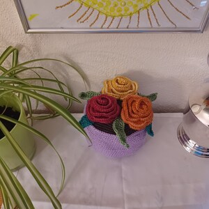 Könnte beinhalten: Eine gehäkelte Blumenarrangements in einem lila Topf. Die Blumen sind rot, orange und gelb. Das Arrangement steht auf einer weißen Oberfläche.