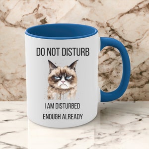 May include: White ceramic coffee mug with a blue rim and handle. The mug features a grumpy cat illustration with the text "Do Not Disturb I Am Disturbed Enough Already."