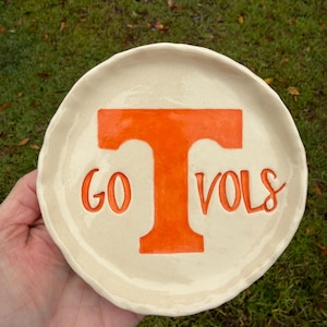 May include: A round, off-white ceramic dish with a scalloped edge. The center features a large, bold orange letter T with the text GO VOLS embossed in orange lettering below the crossbar of the T, creating a clear and simple design.