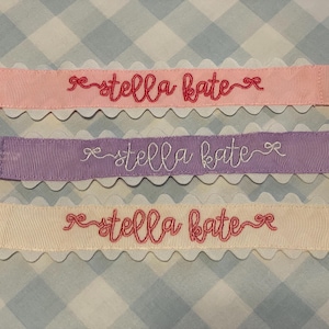 May include: Three pacifier clips featuring ribbed fabric straps in light pink, lavender, and cream. Each strap has a white scalloped edge, a metal clip at one end, and a white elastic loop at the other. The text "stella kate" is embroidered in cursive.