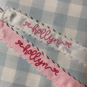 May include: Two white satin ribbon with scalloped edges and pink embroidery. The ribbon is embroidered with the name "Holly" in cursive.