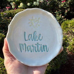 May include: A round, off-white ceramic trinket dish with uneven, wavy edges. The center features a pale yellow sun icon above the teal-colored text "Lake Martin" written in a cursive script. The surface has a glossy, smooth finish.