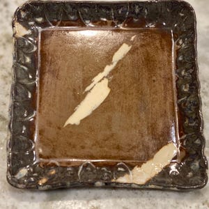 May include: A square ceramic dish with a brown glaze and a white glaze design. The dish has a decorative, scalloped edge.
