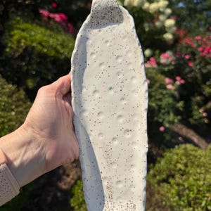 May include: An elongated, oval-shaped ceramic tray with a glossy white glaze and a speckled finish. The surface features a series of small, circular indentations arranged in a pattern, with a textured, speckled handle at each end.