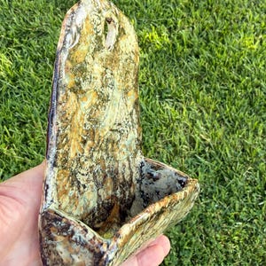 May include: A wall-mounted ceramic planter featuring a mottled glaze in shades of yellow, brown, and dark green. The piece has a tall, arched back panel with a single hole for hanging and a deep, curved basin for holding small items or plants.