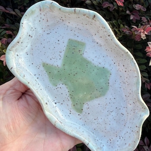 May include: An oval-shaped ceramic trinket dish with an irregular, wavy edge. The surface is off-white with brown speckles and features a smooth, light green silhouette of the state of Texas centered in the middle of the dish.
