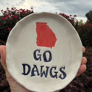 May include: A round, off-white ceramic dish with a hand-painted red silhouette of the state of Georgia. Below the state shape, the text "GO DAWGS" is painted in dark blue, bold, capital letters. The dish has a slightly uneven, organic rim.