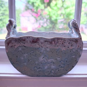 May include: A semi-circular ceramic planter with a light blue glaze, featuring a speckled finish and embossed floral patterns. The rim has a wavy, textured edge with a darker, brownish-purple accent, and two small handles on the sides.