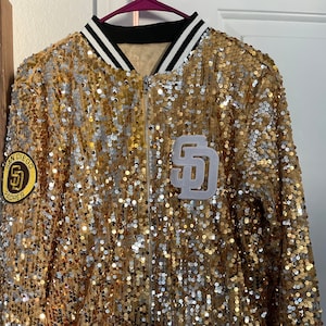 May include: Gold sequin bomber jacket with black and white striped trim. Features a San Diego Padres patch and a white "SD" logo. The jacket has a zipper closure and a black collar with white stripes.