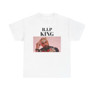May include: White t-shirt with the text "R.I.P KING" above an image of a man wearing a crown and jewellery. The man in the image is wearing a black shirt and has a beard. The background is a soft pink.