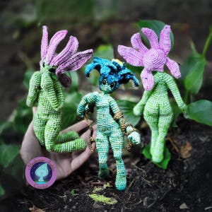 Könnte beinhalten: Drei handgefertigte Häkelfiguren. Zwei Figuren haben grüne Körper und lila blütenförmige Köpfe. Die dritte Figur hat einen blau-grünen Körper und einen blauen Kopf. Die Figuren sind in einer natürlichen Umgebung posiert.