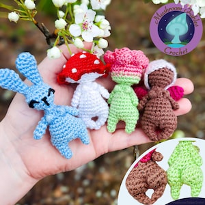 Op de afbeelding: Een verzameling handgemaakte gehaakte figuren, waaronder een blauw konijn, een paddenstoel met een rode dop en verschillende grillige karakters met bloemachtige hoofden. De figuren zijn in verschillende kleuren en worden tentoongesteld tegen een natuurlijke achtergrond.