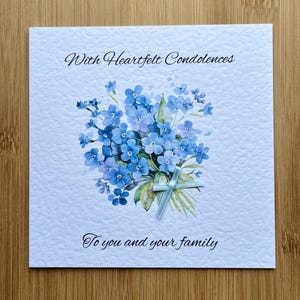 May include: A sympathy card featuring a watercolour illustration of blue forget-me-not flowers. The card is inscribed with "With Heartfelt Condolences" and "To you and your family". A light blue ribbon is tied around the floral arrangement.