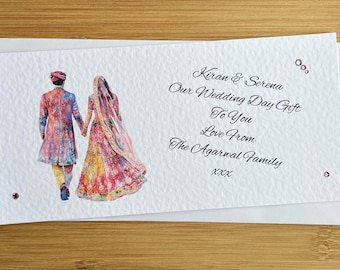 May include: A white wedding card with a watercolor illustration of a couple in traditional Indian attire. The card includes the text "Kiran & Serena, Our Wedding Day Gift To You, Love From The Agarwal Family, xxx."