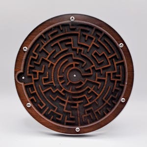 Pode incluir: Um jogo de labirinto circular de madeira com uma moldura de madeira marrom escura e um design de labirinto preto. O labirinto tem uma pequena bola de metal no centro.