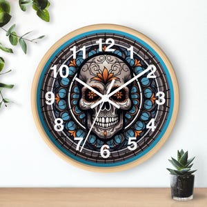 May include: Round wall clock with a light wood frame, featuring a detailed skull design in the center. The skull has intricate floral patterns in orange and white, set against a blue and gray background. White numerals mark the hours.