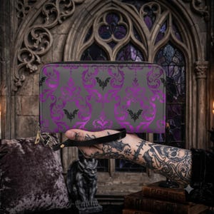 May include: A rectangular zip-around wallet featuring a dark gray base with a repeating purple damask pattern and small black bat silhouettes. The wallet includes a black wrist strap attached to a silver-toned metal zipper pull and hardware.