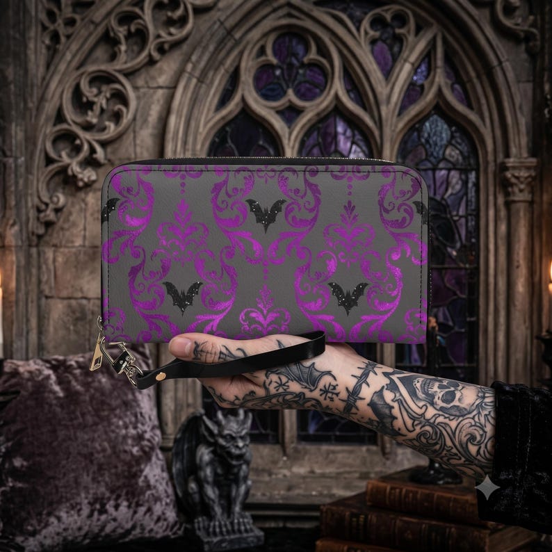 May include: A rectangular zip-around wallet featuring a dark gray base with a repeating purple damask pattern and small black bat silhouettes. The wallet includes a black wrist strap attached to a silver-toned metal zipper pull and hardware.