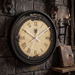 May include: A round wall clock with a black frame and a beige face featuring Roman numerals. A dragon encircles the dial, with its head at the top and wings on the sides. A small shield emblem is centered below the twelve o'clock position.