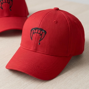 May include: A red baseball cap with a curved brim. The cap features a black embroidered design of vampire fangs with a drop of blood. The cap is made of a durable fabric and has a structured fit.