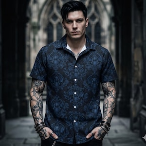May include: Dark navy short-sleeve button-up shirt with a repeating bat and scrollwork pattern in a lighter blue. The shirt has a white collar and is worn by a person with tattoos and jewelry.