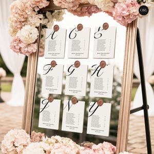 May include: A framed seating chart with a gold-colored frame, featuring individual cards labeled with letters A through Y. Each card has a list of names and is sealed with a wax stamp. The frame is decorated with pink and white flowers.