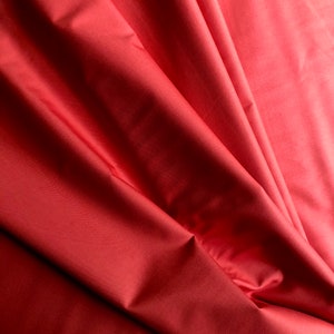 Könnte beinhalten: Eine Nahaufnahme eines gefalteten Stoffstücks in einem tiefen Rot. Der Stoff hat eine glatte, seidige Textur und ist so gefaltet, dass er eine fächerartige Form bildet.