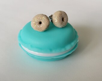 May include: A pair of donut-shaped earrings with glitter accents, resting on a turquoise and white macaron-shaped container. The earrings are beige with a sparkly coating, and the container is a bright, solid color.