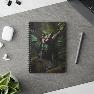 Op de afbeelding: Een spiraalgebonden notitieboek met een groene en bruine afbeelding van een wezen met vleugels en een harige lichaam. Het wezen heeft een groen en bruin gepantserd borststuk en zit op een rots in een bos.