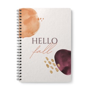 Può includere: Un quaderno a spirale con una copertina bianca che presenta disegni astratti ad acquerello in tonalità di arancione, bordeaux e oro. Il testo "HELLO fall" è stampato in nero e arancione sulla copertina.