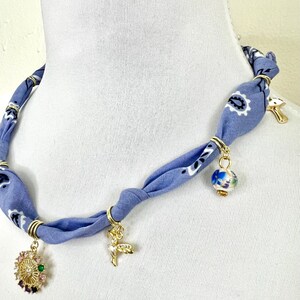 May include: A blue bandana necklace with gold-colored metal rings and charms. The charms include a gold-colored deer, a gold-colored mushroom, a colorful floral pendant, and a blue and white globe pendant.