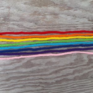 May include: A set of ten crocheted rainbow-colored snakes with black eyes. The snakes are arranged in a row from red to pink, with each snake slightly shorter than the one before it.